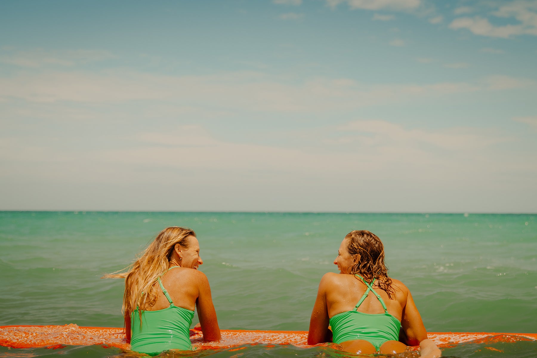 Two women in green swimsuits hanging on an orange surfboard in the ocean with a clear sky.
